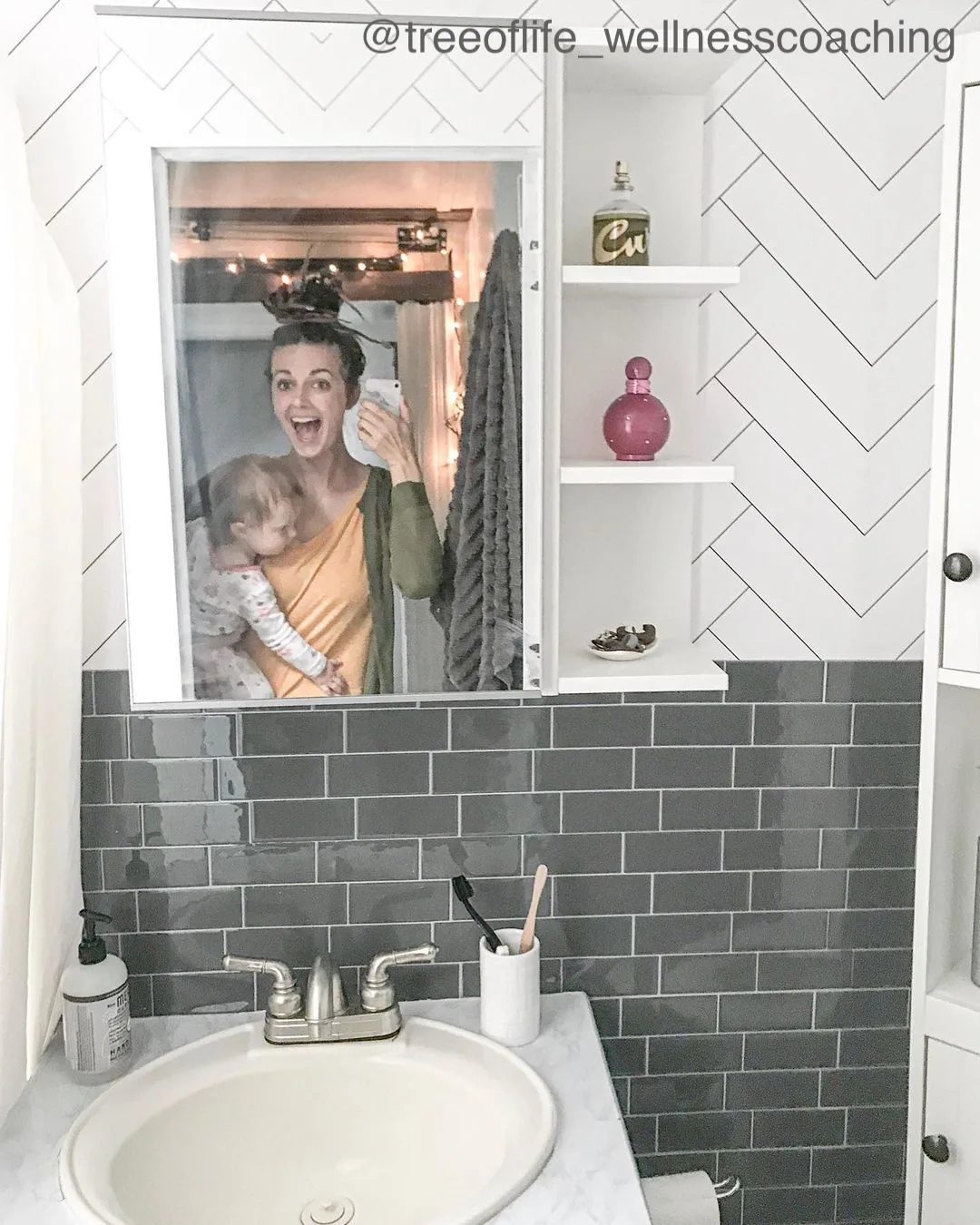 Smiling woman seen holding a baby in the reflection of a small bathrooms mirror.